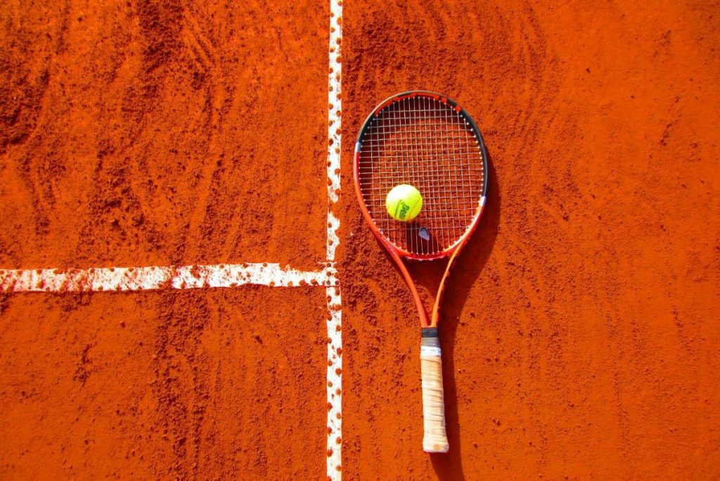 Raqueta de tenis acostada sobre suelo de cancha de arcilla con una pelota de tenis encima.